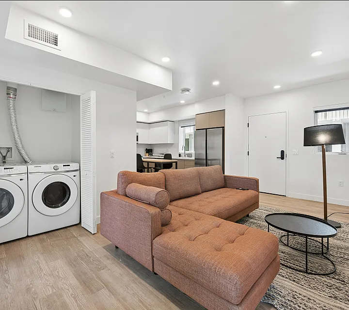 Modern living area showcasing a stylish orange sofa, open kitchen with stainless steel appliances, and convenient in-unit laundry space.