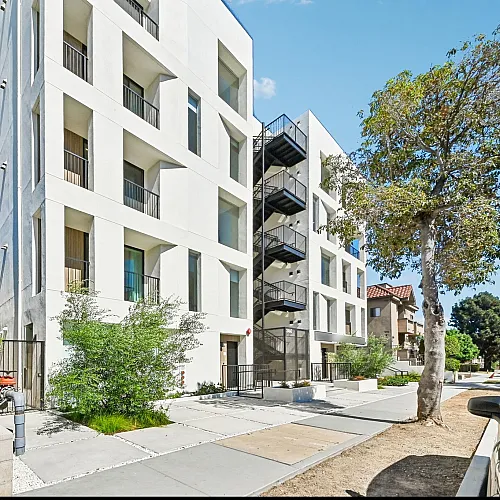 Contemporary apartment building with a sleek white facade, large windows, and surrounding lush greenery on a sunny day.