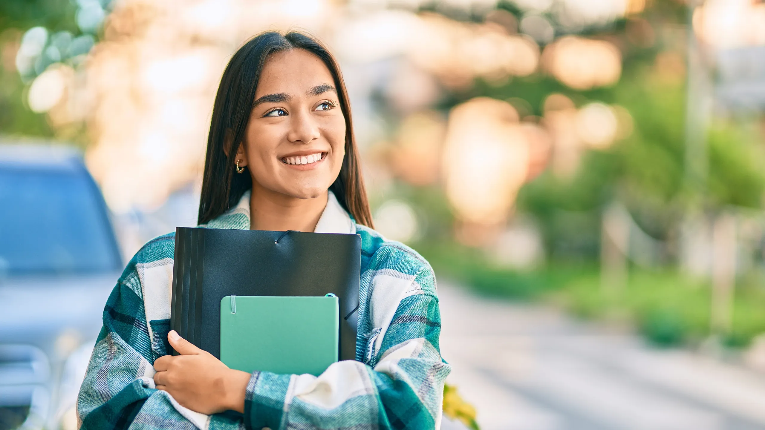 Confident young woman with long hair, smiling brightly while holding folders outdoors, exuding positivity and success.