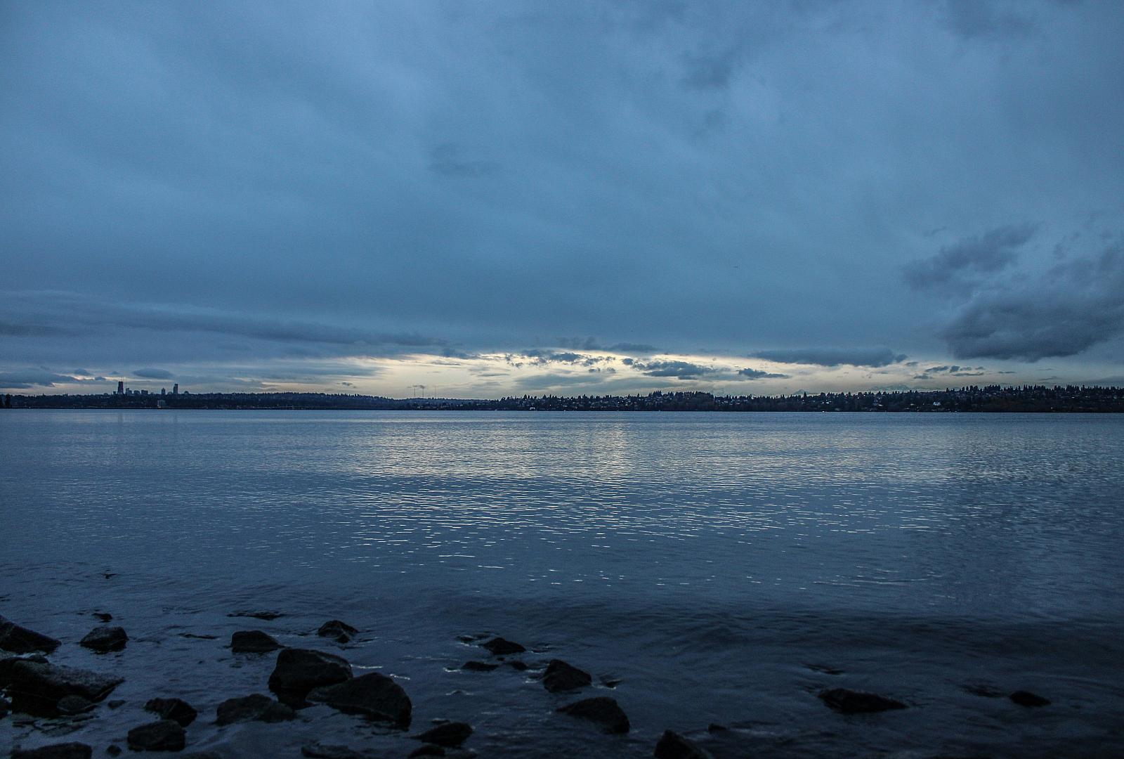 Serene view of a tranquil lake under a dramatic cloudy sky at dusk, with a silhouette of a distant city skyline on the horizon.