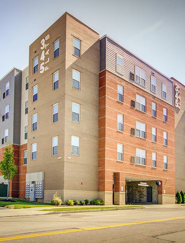 Modern multi-story apartment building with a blend of brick and grey facade, featuring multiple windows and green landscaping.