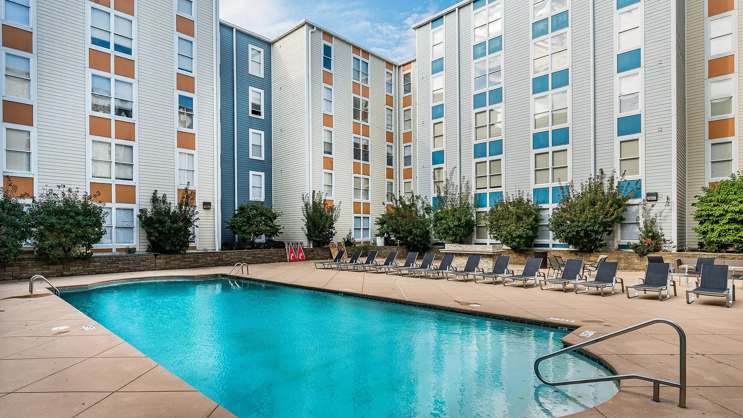 Sunny courtyard view featuring a sparkling blue pool surrounded by modern apartment buildings, with ample lounge chairs for relaxation.