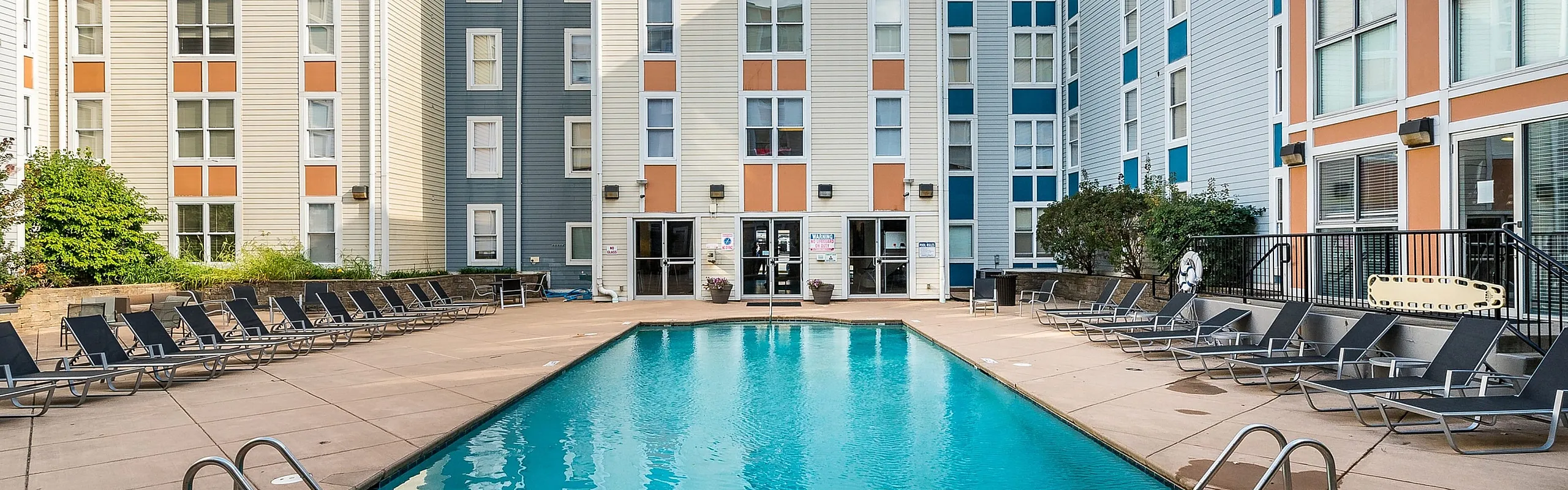 Chic urban apartment complex featuring a sparkling outdoor pool with sleek lounge chairs, surrounded by vibrant, multi-story residential buildings.