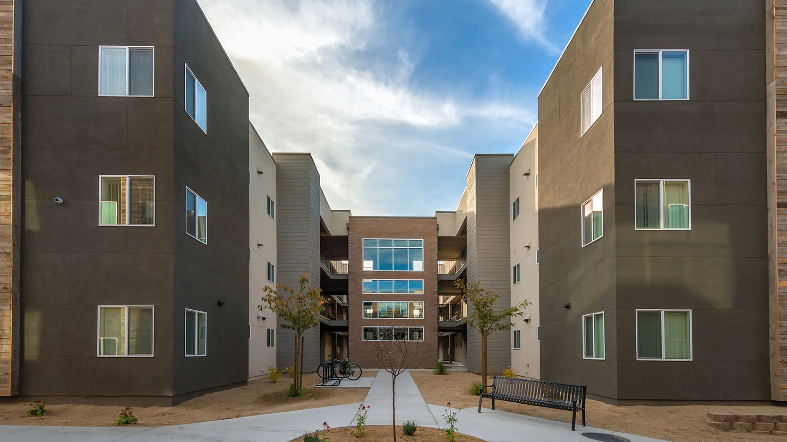 Modern apartment complex with sleek architecture, featuring a central courtyard with landscaped paths and seating under a vibrant sky.