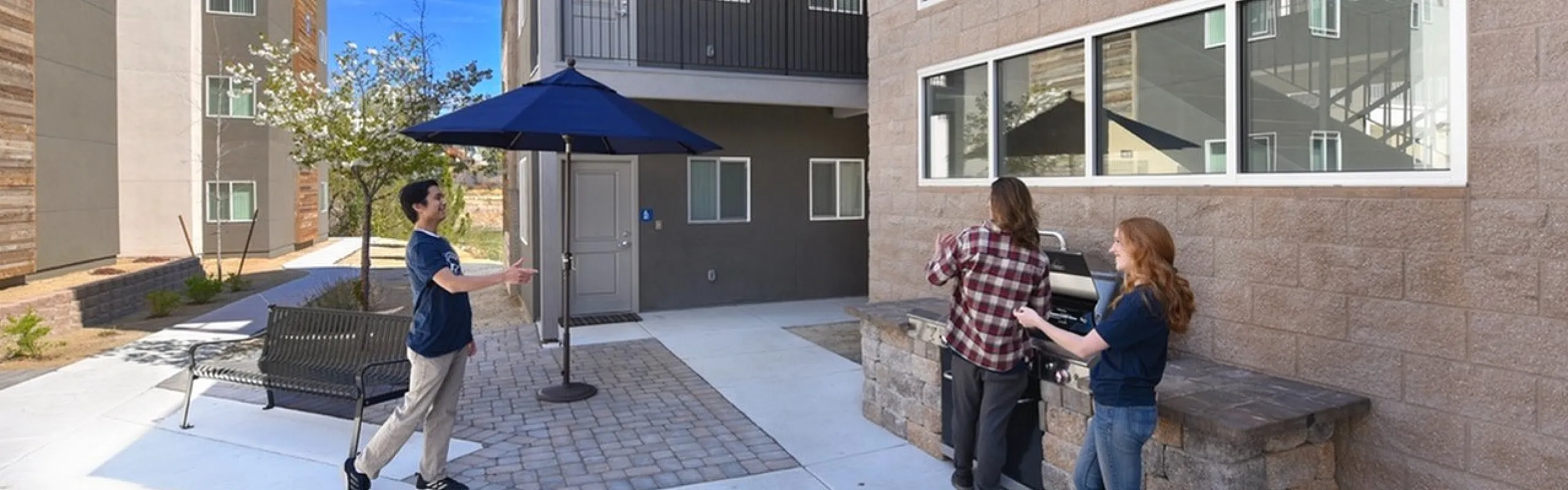 Modern apartment courtyard with a vibrant social scene, featuring a brick barbecue area, stylish outdoor seating, and beautiful landscaping under a bright blue sky.