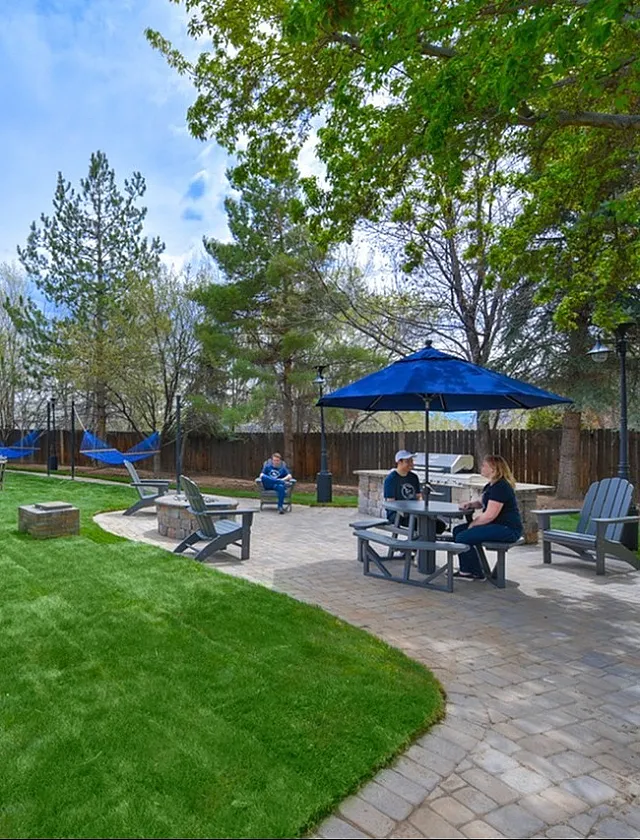 Inviting outdoor patio with green lawn, picnic tables, and blue hammocks set under vibrant trees, creating a perfect space for relaxation and socializing.