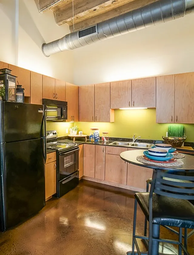 Modern kitchen with sleek black appliances, warm wooden cabinetry, and a cozy dining area, featuring industrial-chic exposed ductwork and polished concrete floors.