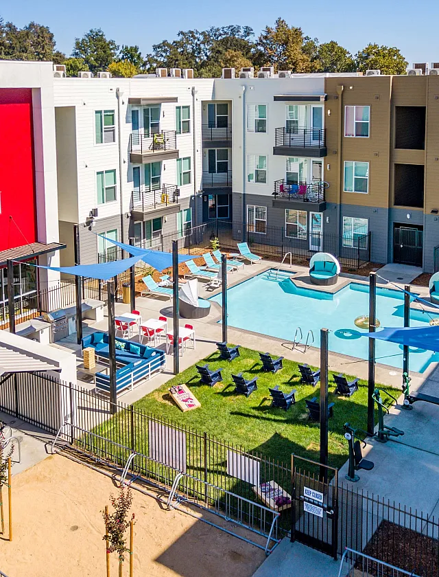 Vibrant apartment complex featuring a modern pool area with stylish cabanas, shaded seating, and colorful facade under a clear blue sky.