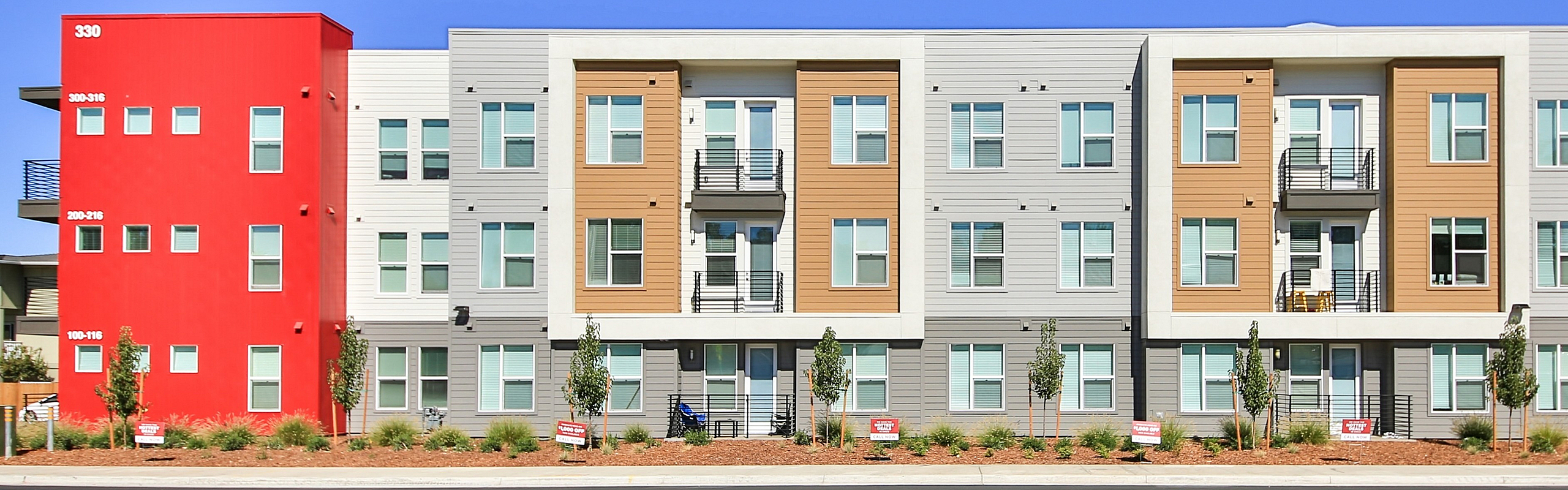 Modern apartment complex with vibrant red accent wall, spacious balconies, and lush landscaping.