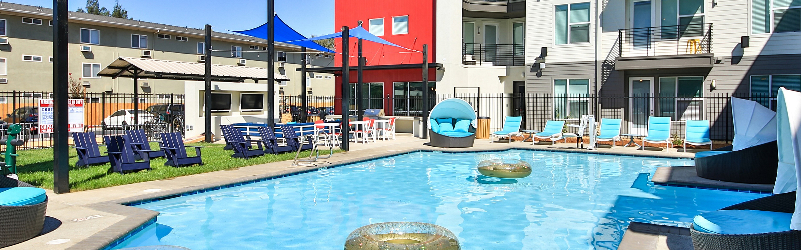 Vibrant apartment complex courtyard featuring a sparkling blue swimming pool with lounge chairs and modern architecture under sunny skies.
