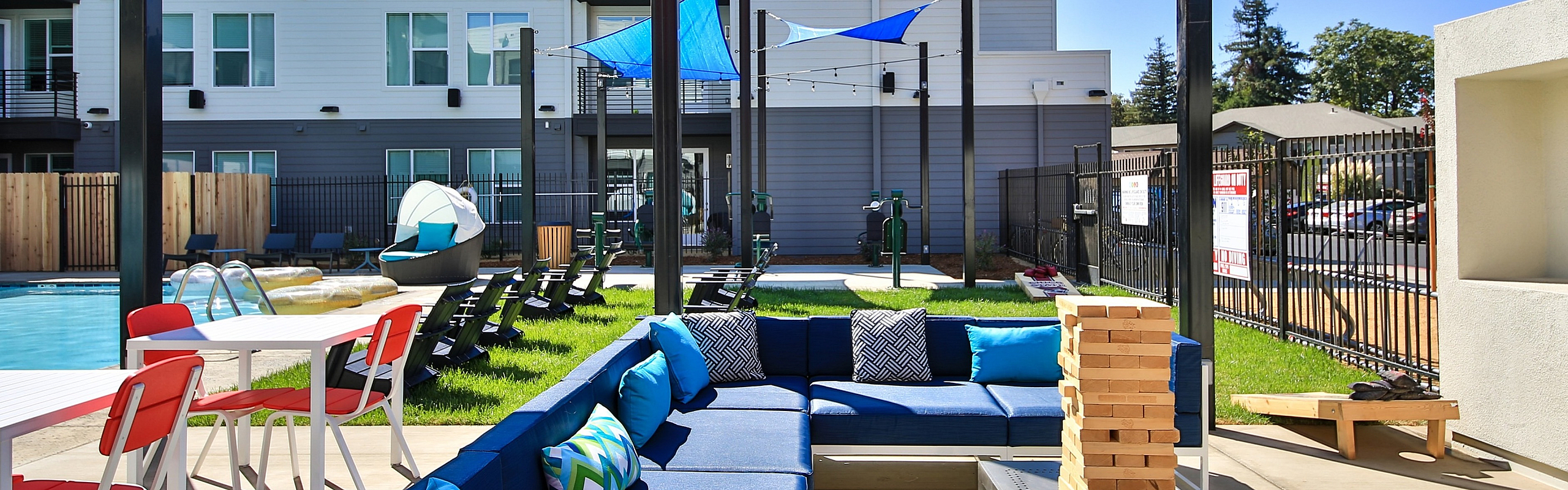Modern outdoor community space with stylish blue seating, dining tables, vibrant sunshades, and a refreshing pool in a contemporary apartment complex.