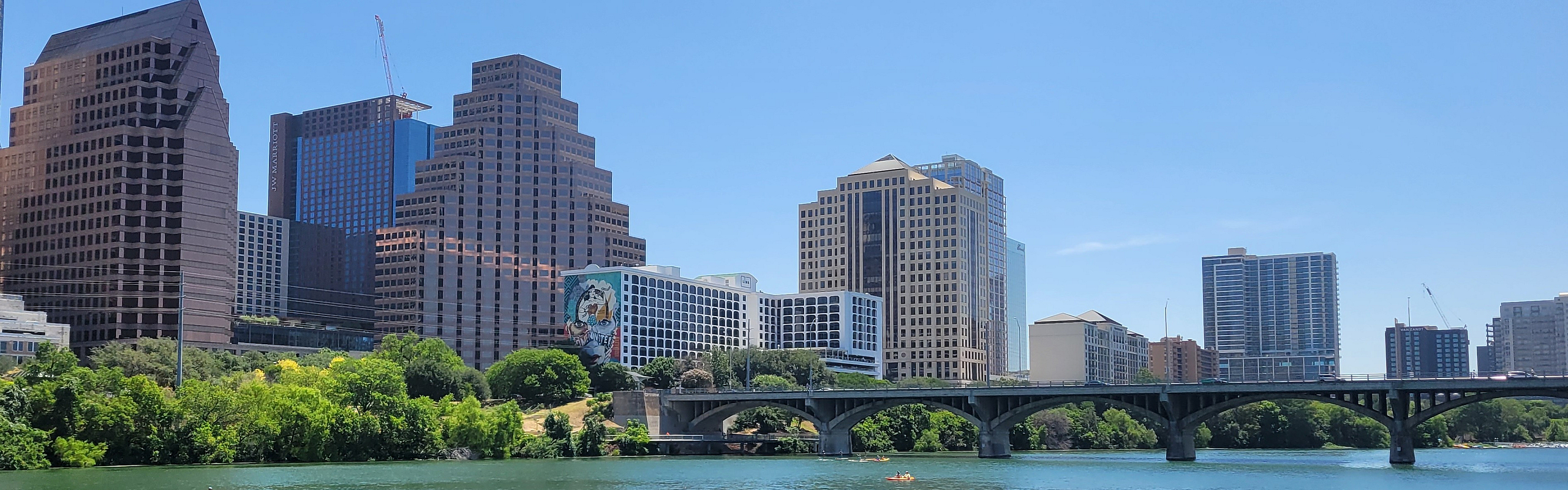 Scenic riverside skyline featuring modern skyscrapers, lush greenery, and lively water activities under a clear blue sky.