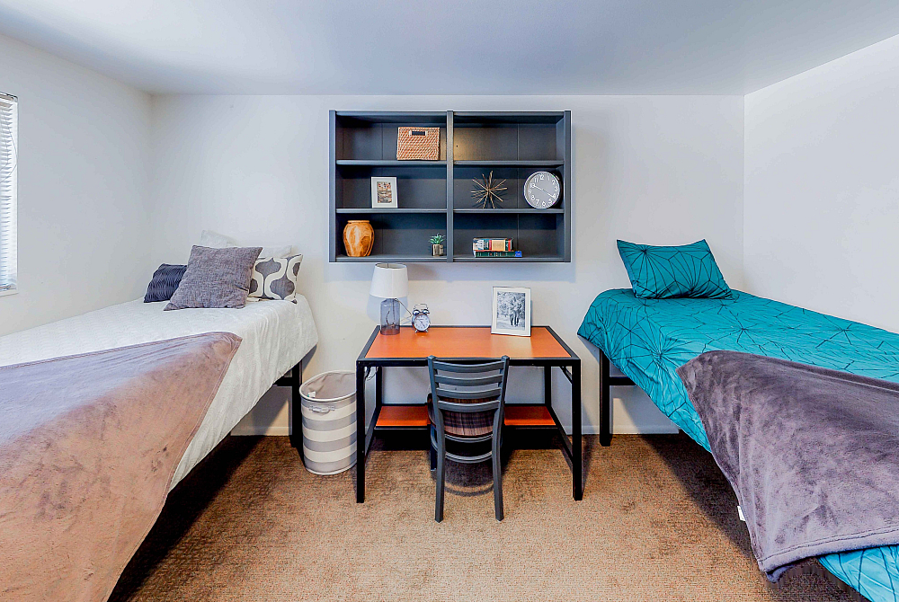 Cozy dual-bedroom setup featuring two twin beds, modern shelving with stylish decor, and a compact study desk by a sunlit window.