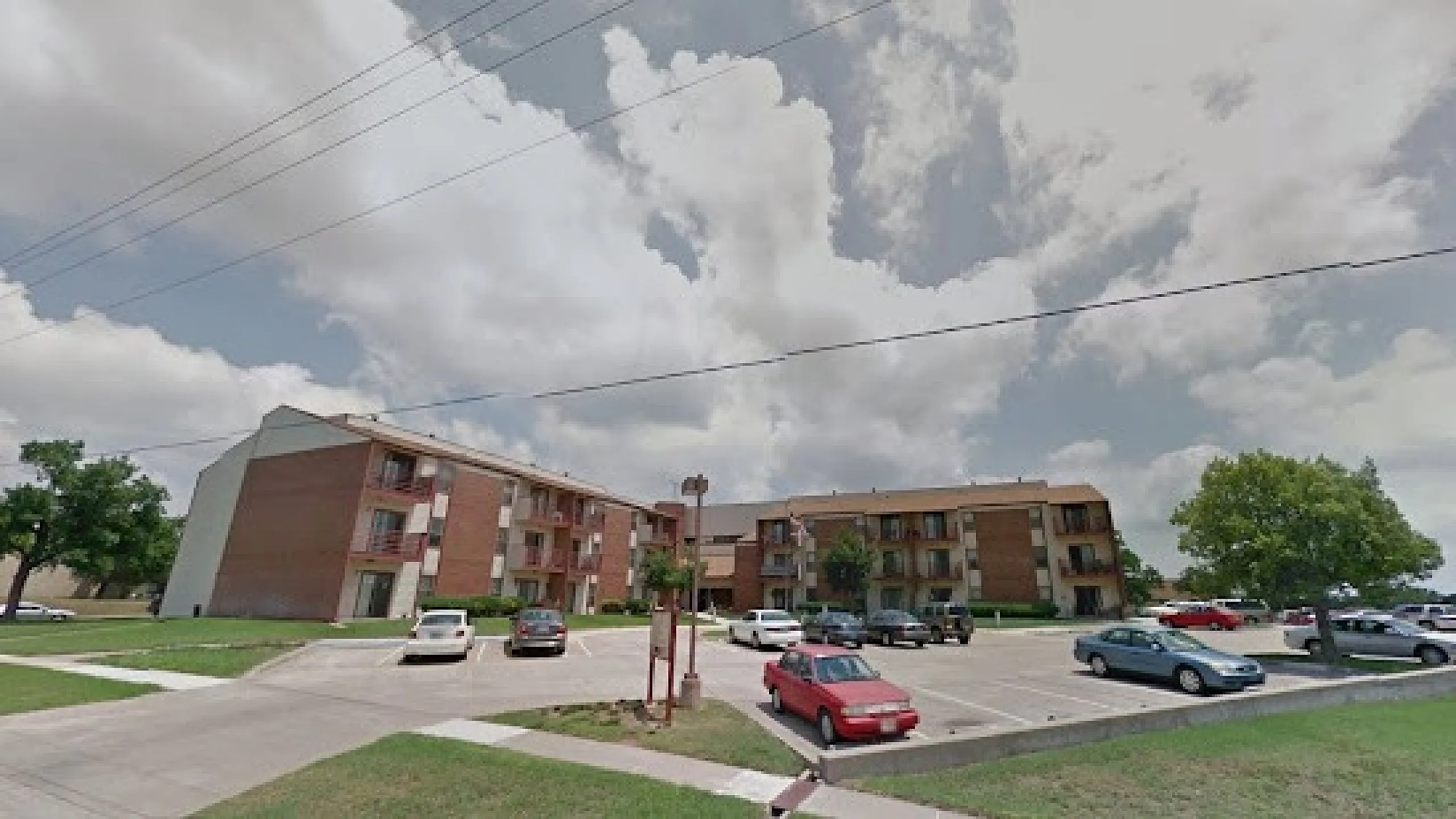 Lovely apartment complex with ample parking and lush greenery under a bright, cloudy sky.