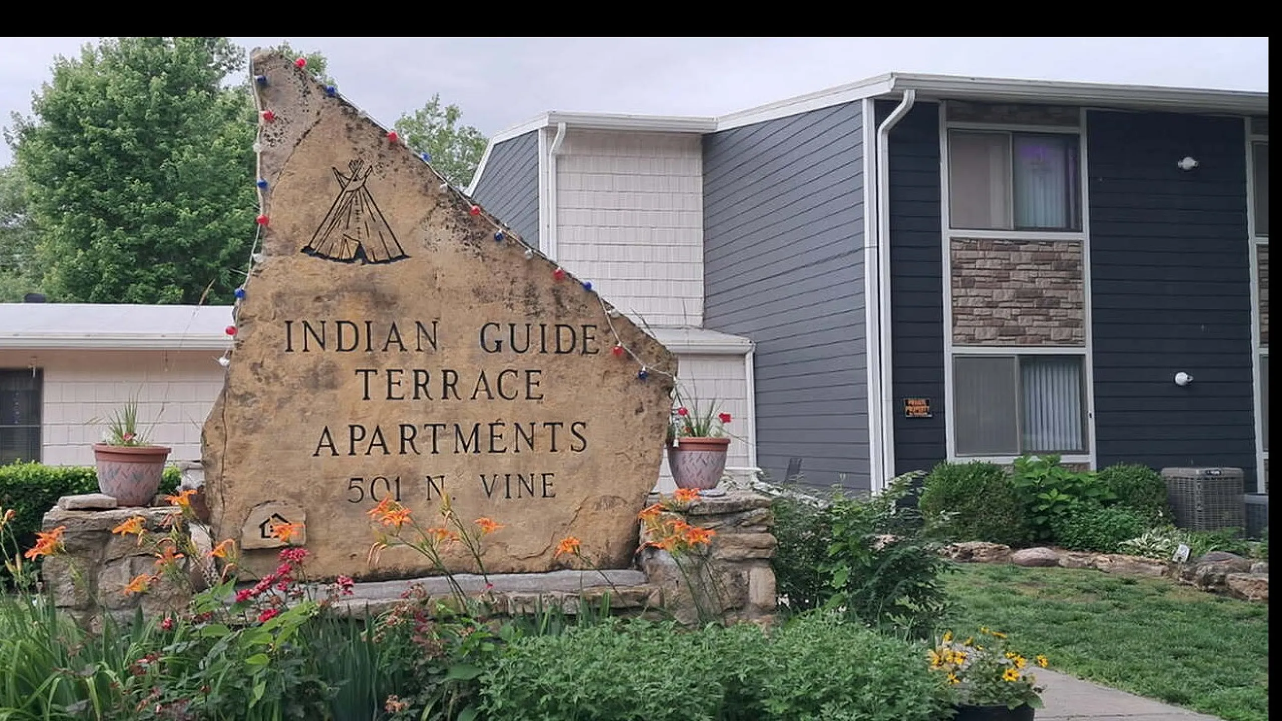 Charming entrance to Indian Guide Terrace Apartments with a rustic stone sign surrounded by vibrant garden flowers.