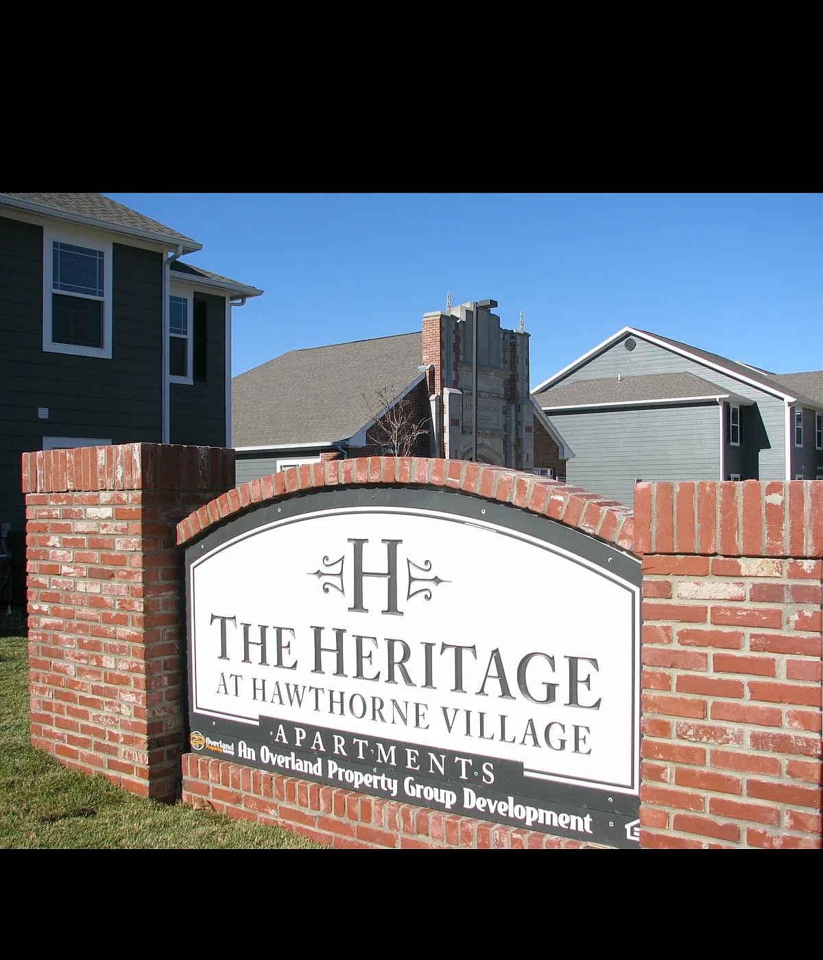 Charming entrance to The Heritage at Hawthorne Village Apartments, showcasing classic brick architecture against a clear blue sky.