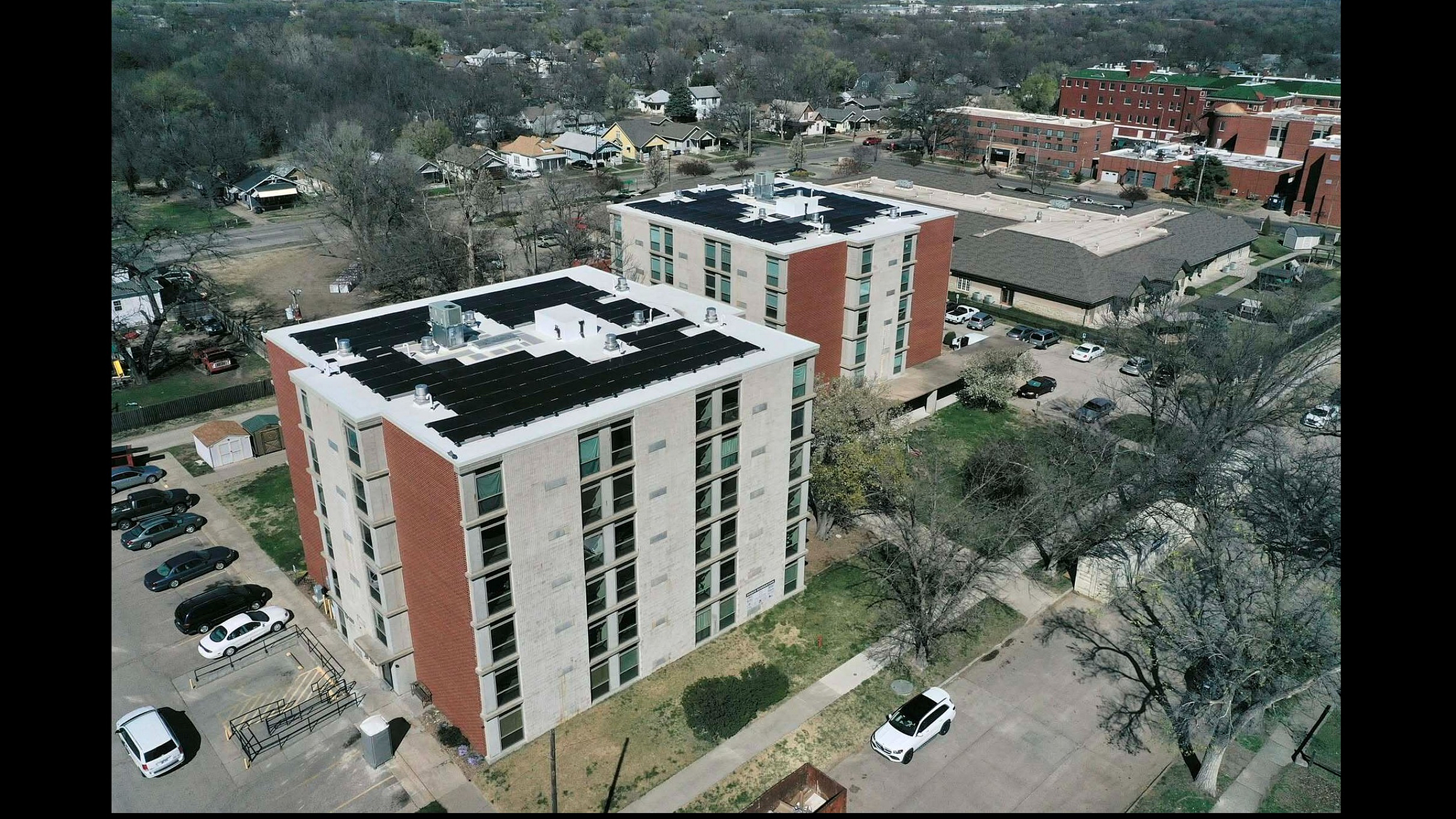 Aerial view of stylish modern apartment buildings with solar panels and ample parking, surrounded by lush greenery in a tranquil neighborhood setting.