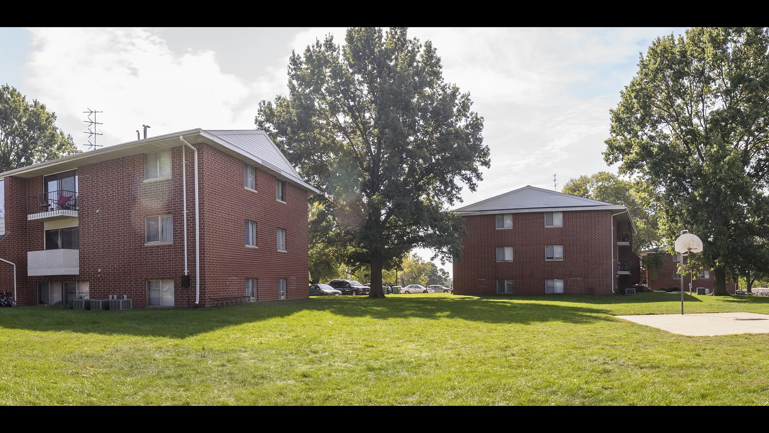 Charming brick apartment buildings surrounded by lush green lawns and mature trees, featuring an outdoor basketball court.