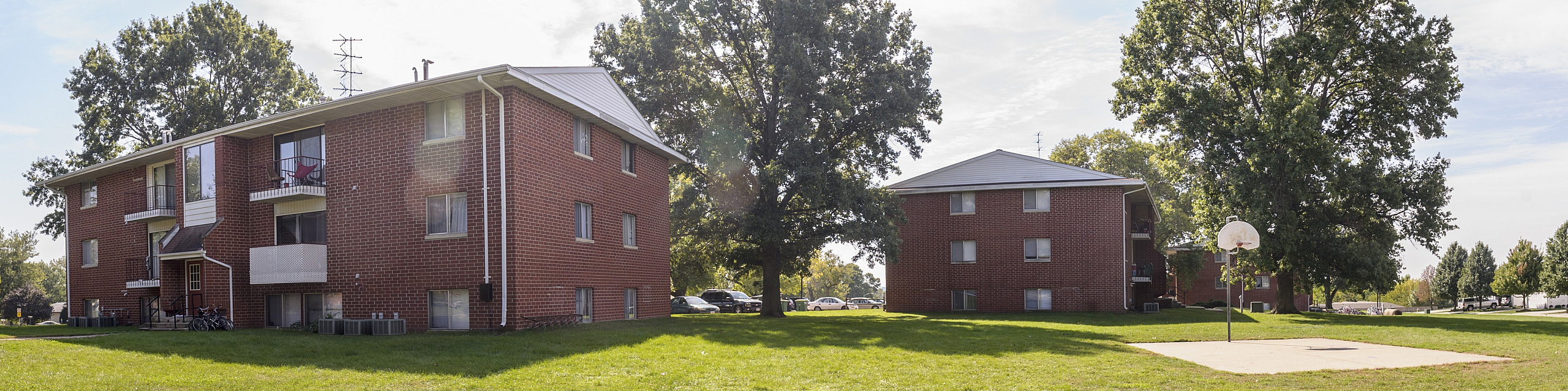 Inviting red brick apartment buildings set in a lush green landscape with mature trees and a nearby basketball court.