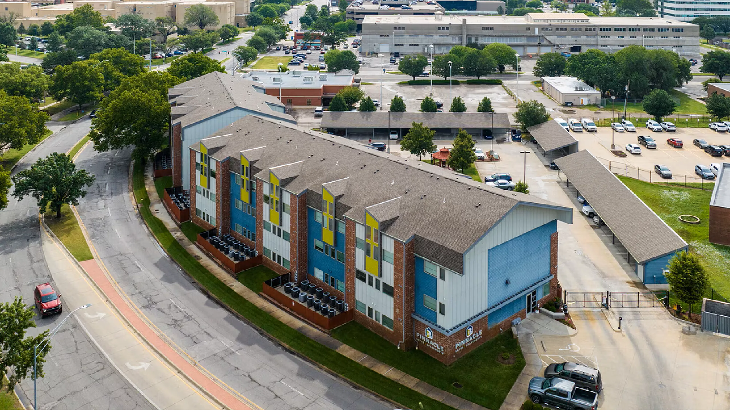 Aerial view of a vibrant apartment complex with modern architecture, ample parking, and lush greenery, set in a bustling urban environment.