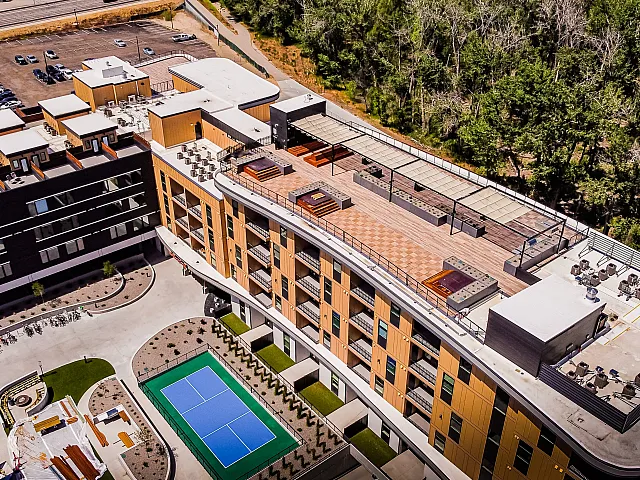 Vibrant apartment complex with spacious rooftop terrace featuring lounge areas, hot tubs, and lush greenery. Includes a modern tennis court and ample parking, set against a stunning natural backdrop.