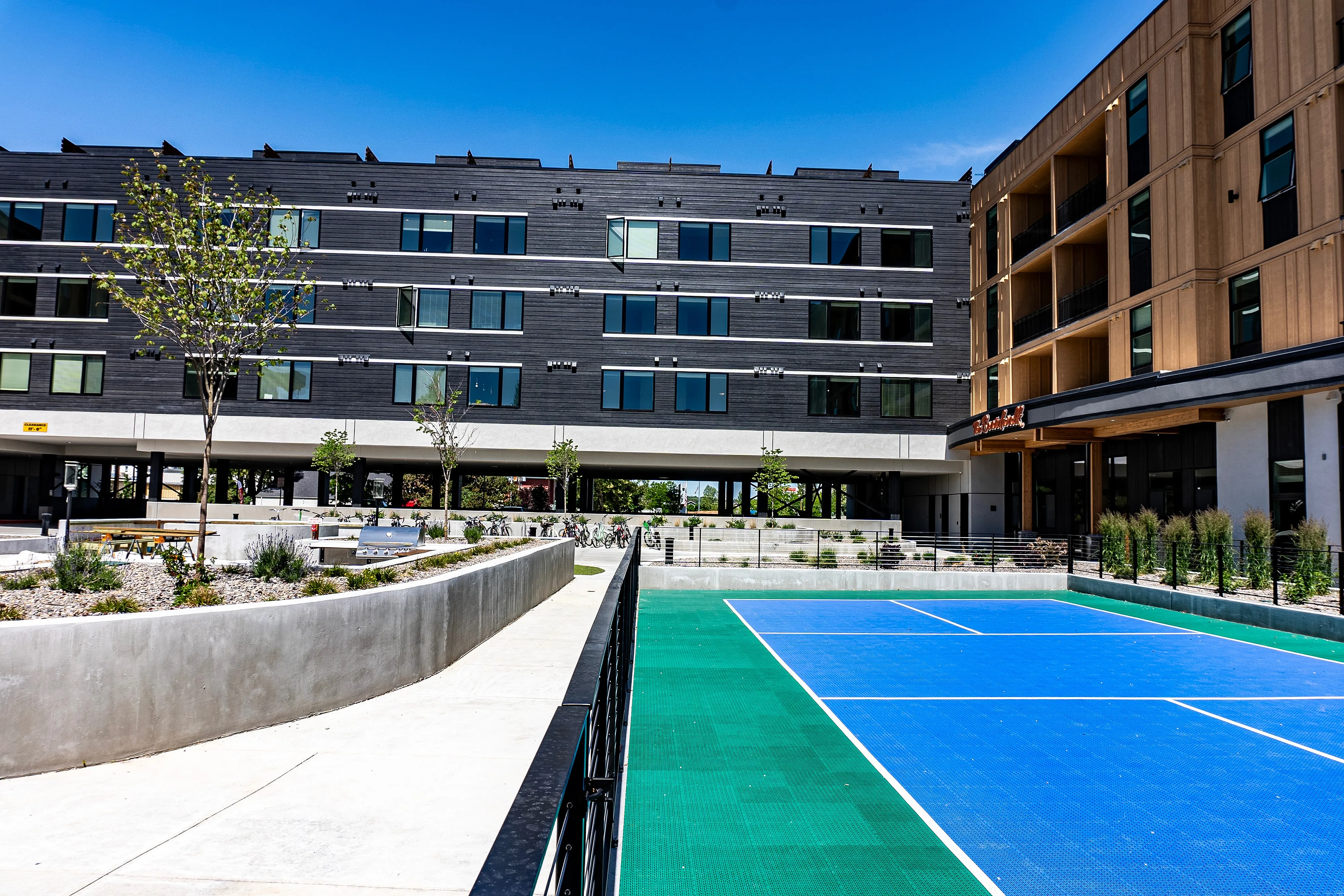 Modern apartment complex with sleek design, showcasing a vibrant blue tennis court and landscaped surroundings under a clear blue sky.