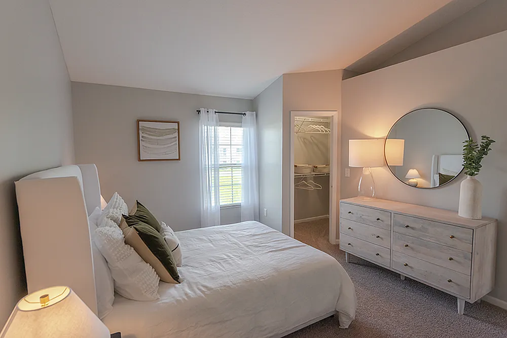 Bright and inviting bedroom with elegant gray walls, cozy bed with decorative pillows, ample natural light from large window, chic dresser with round mirror, and modern lamp.