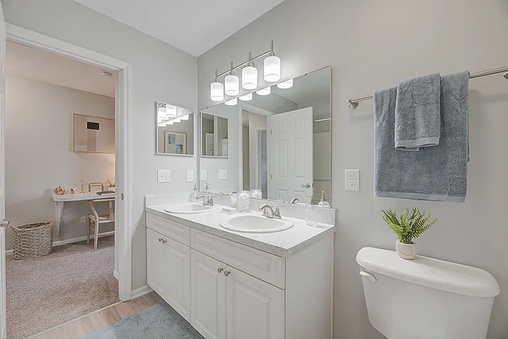 Bright and modern bathroom with double sinks, sleek mirror, and soft blue accents, leading to a cozy office nook.