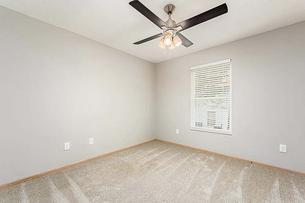 Bright and inviting bedroom with soft carpeting, modern ceiling fan, and a sunlit window offering a serene atmosphere.