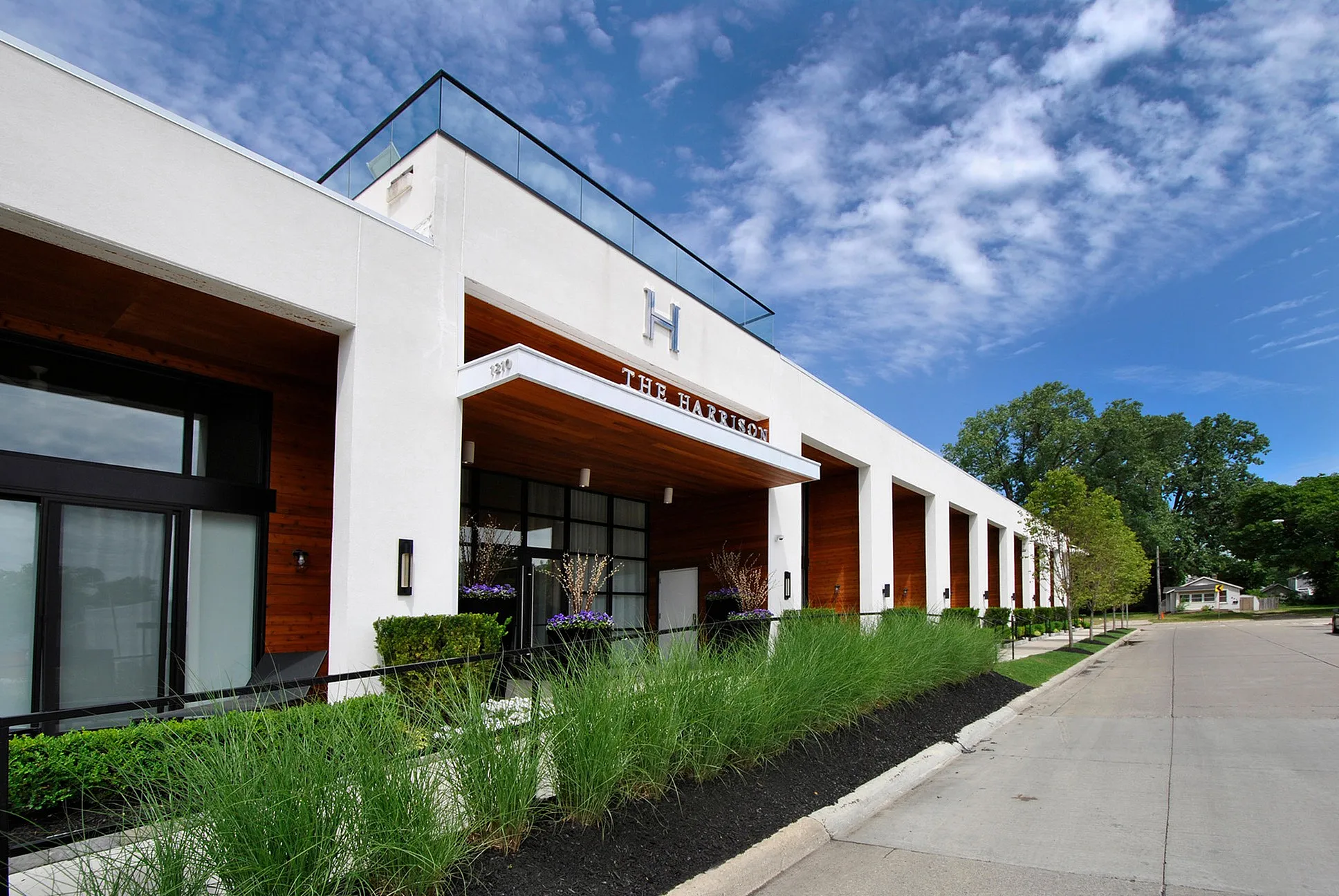 Modern building exterior of The Harrison, featuring sleek architecture with glass railings and lush landscaping against a blue sky backdrop.