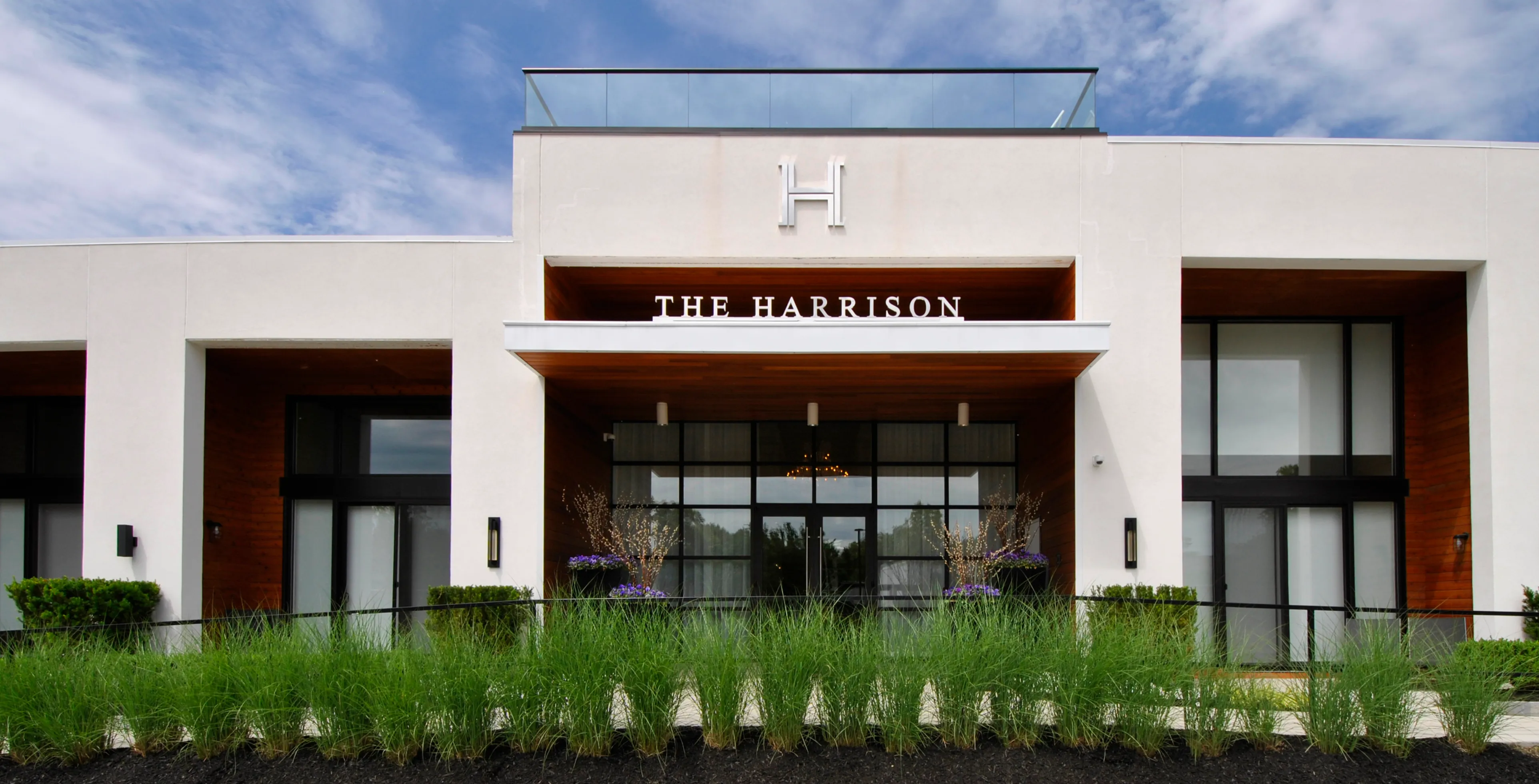 Modern building exterior of The Harrison, featuring sleek architecture with glass railings and lush landscaping against a blue sky backdrop.
