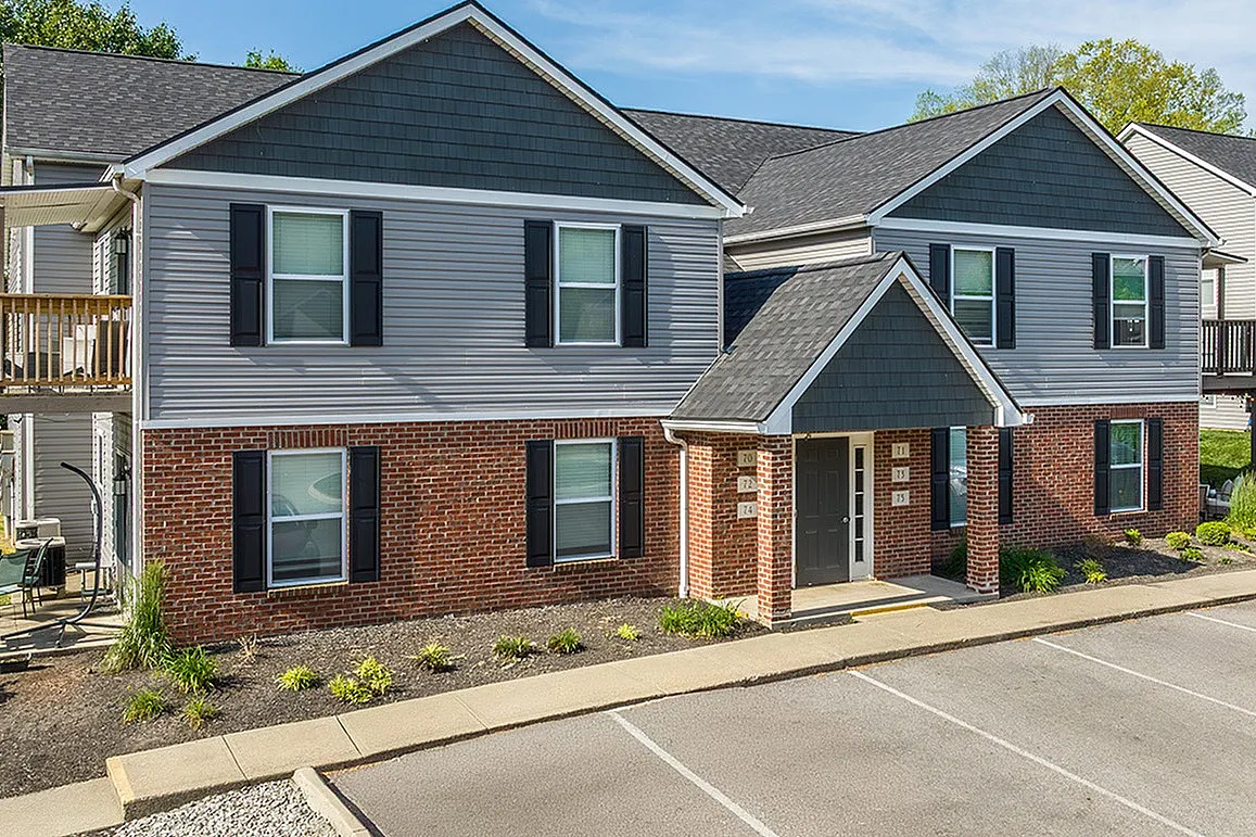 Charming multi-unit apartment building with brick accents, surrounded by lush greenery and featuring a welcoming outdoor patio.