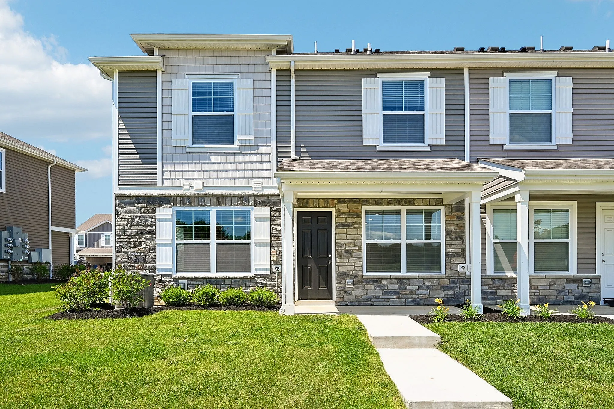 Charming modern townhouse exterior with stone accents, inviting front porch, and lush green lawn under a clear blue sky.