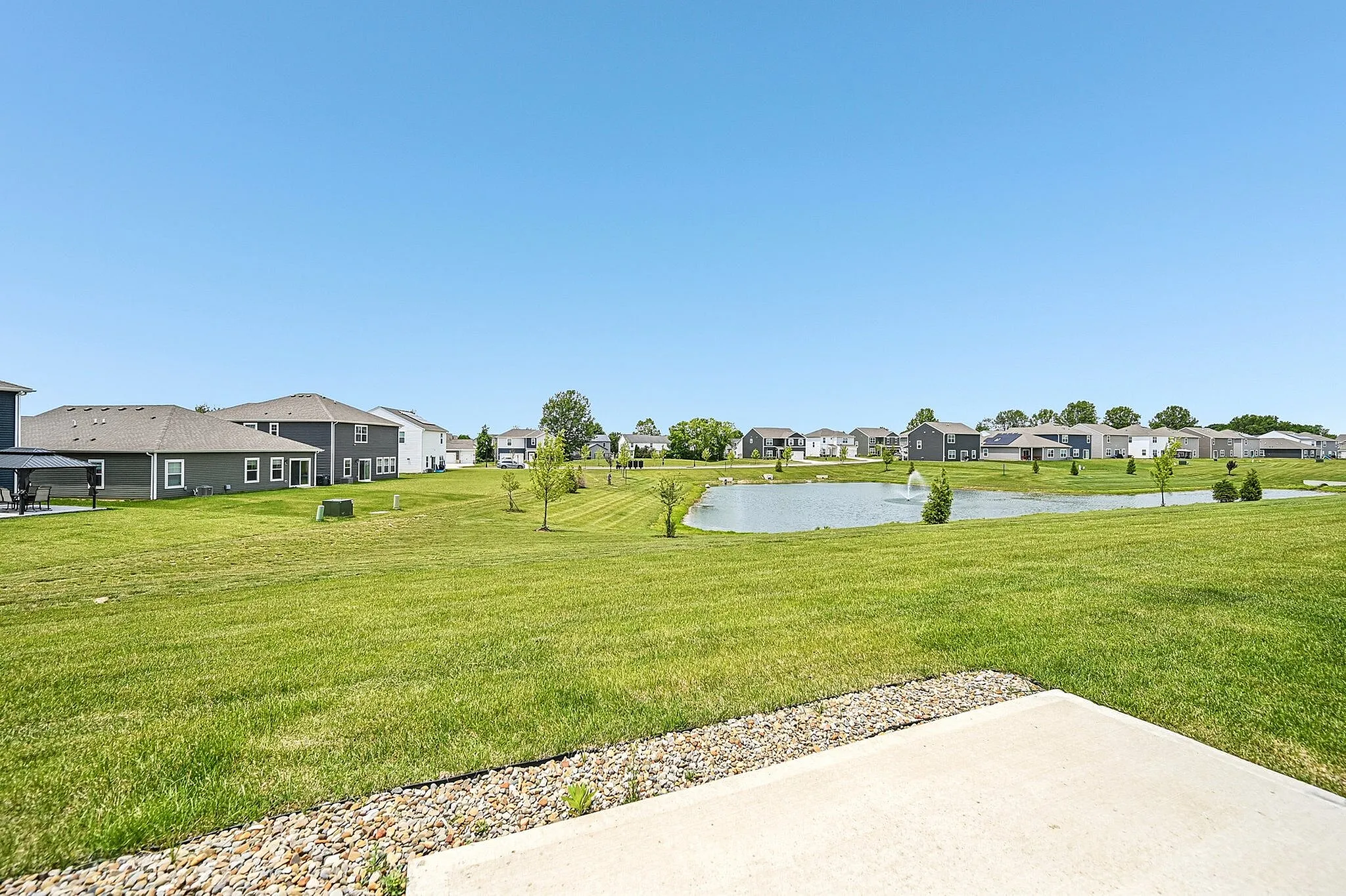 Charming neighborhood scene with lush green lawns, modern homes, and a serene pond under a clear blue sky.