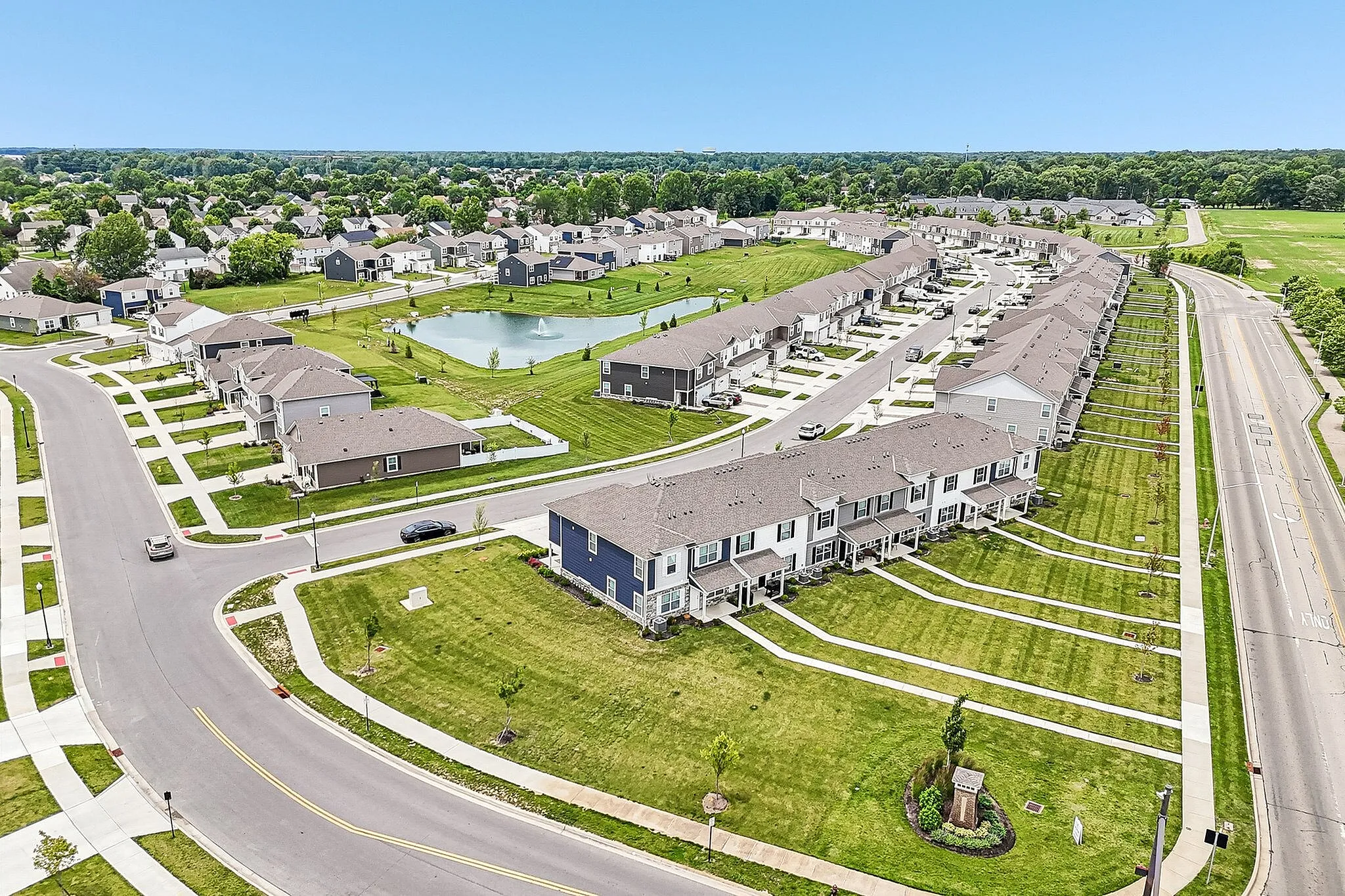 Tranquil suburban community with elegant row homes, lush green lawns, and a serene pond with a fountain, offering a picturesque neighborhood vibe.