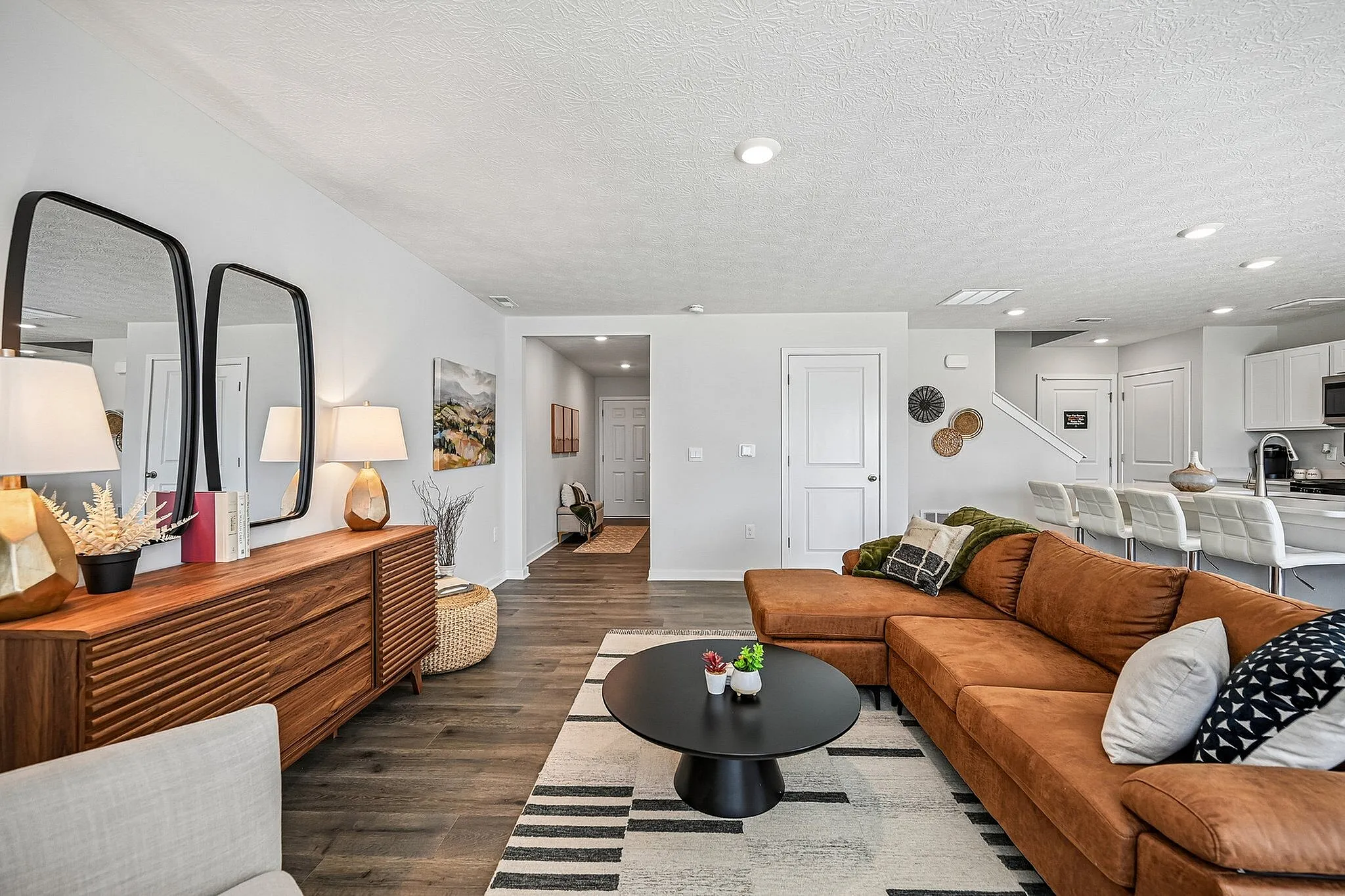 Contemporary living room with stylish decor, featuring a cozy brown sectional, sleek wood console with elegant mirrors, and open kitchen with bar seating.