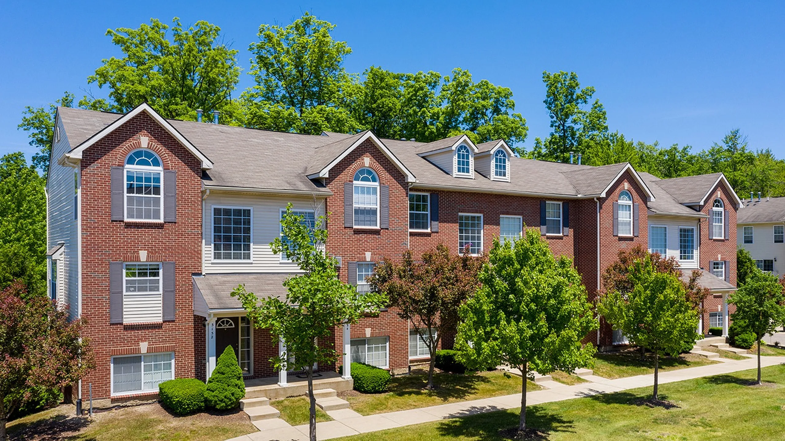 Charming townhouse community with elegant brick facades, lush greenery, and inviting sidewalks under a clear blue sky.