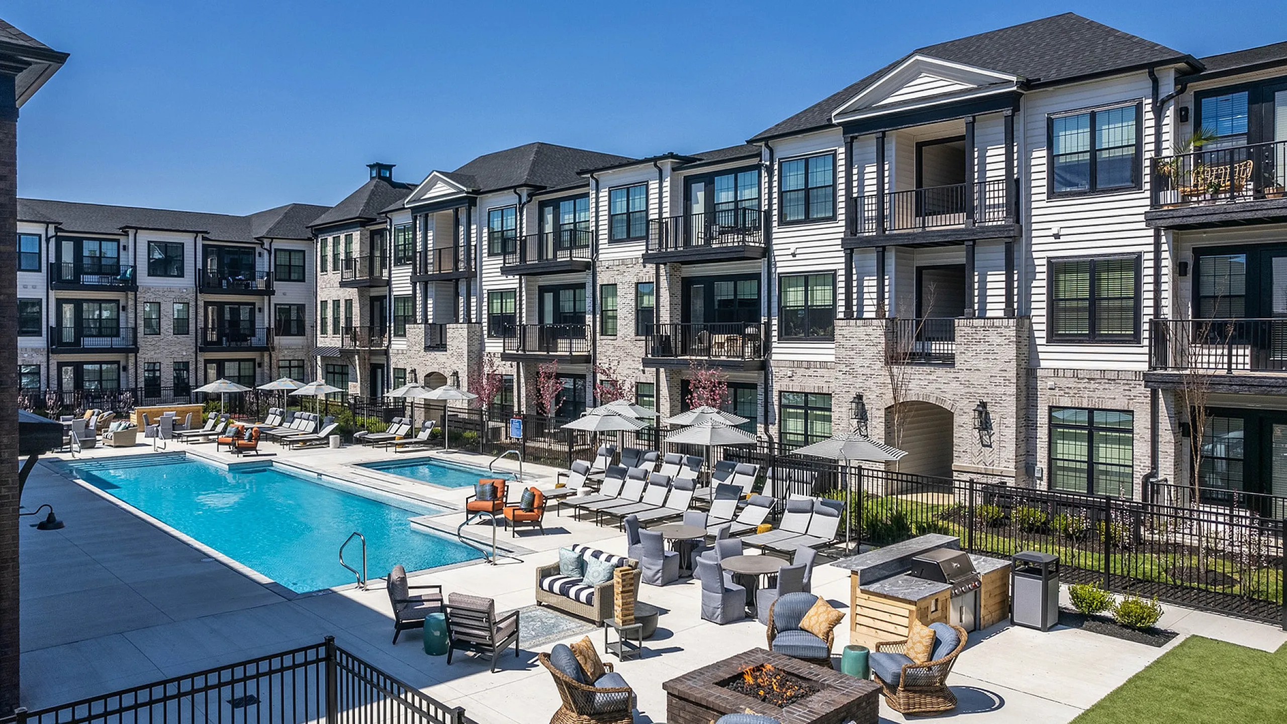 Luxurious apartment complex with elegant architecture, highlighting an inviting swimming pool, spacious sun deck, and cozy fire pit area amid beautifully landscaped surroundings.