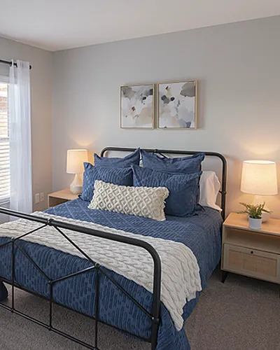 Inviting bedroom with elegant decor, featuring a cozy bed with blue bedding, stylish lamps, and modern artwork, bathed in natural light from large windows.