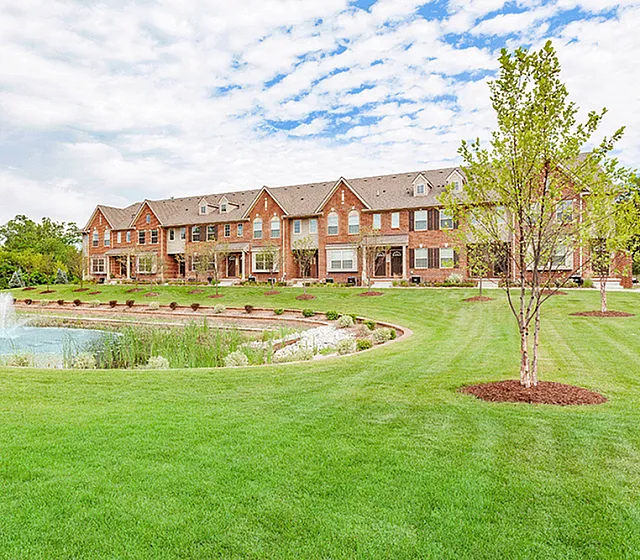 Charming townhouses with rustic brick facade overlooking a serene pond and lush green lawn under a bright blue sky.