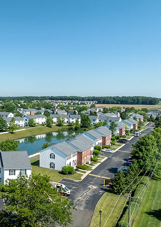 Aerial view of a serene residential community with charming townhouses, lush green spaces, and a tranquil pond under a clear blue sky.