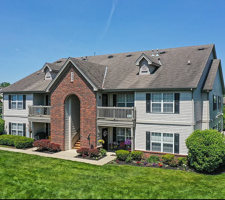 Charming two-story apartment building with welcoming brick archway, surrounded by lush greenery and vibrant flowerbeds under a clear blue sky.
