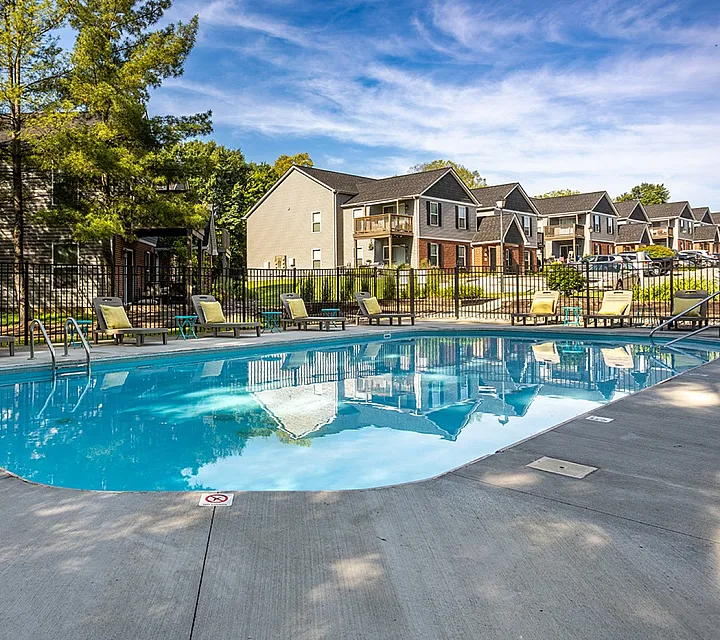Inviting apartment community showcasing a sparkling pool surrounded by lounge chairs, adjacent to charming townhomes under a clear blue sky.