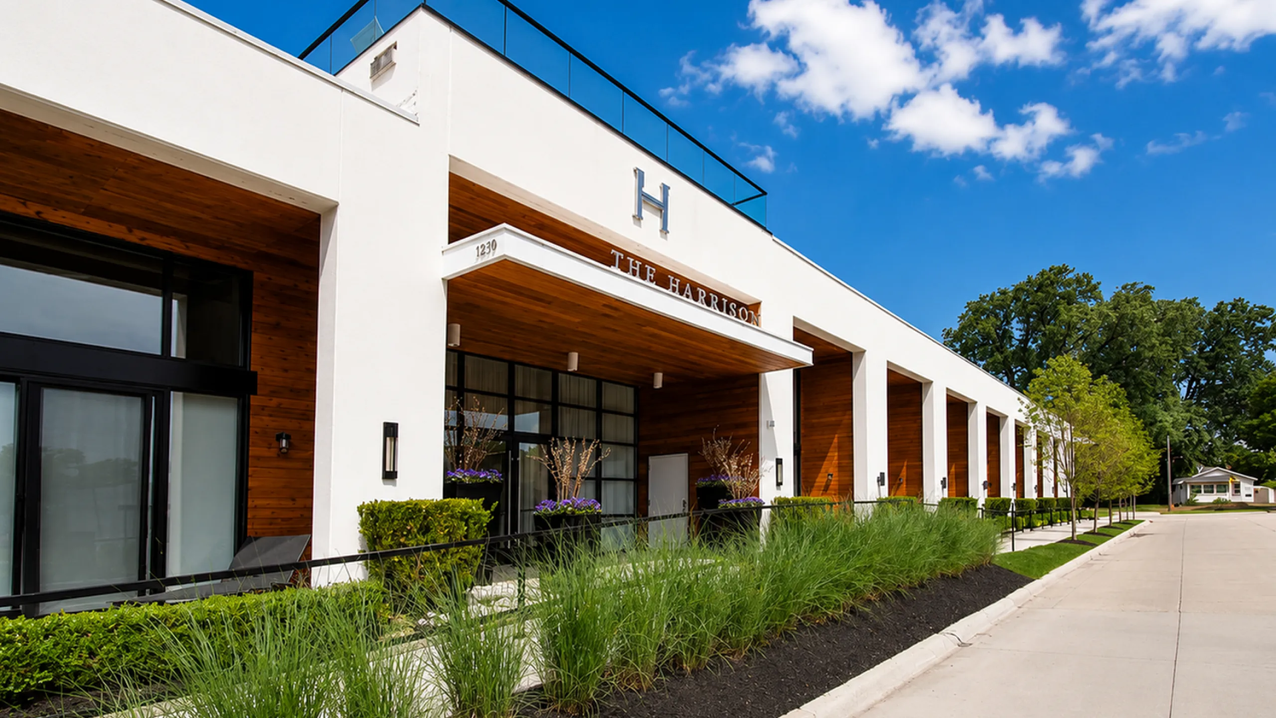 Modern luxury apartment exterior of The Harrison, showcasing sleek architecture with natural wood accents and vibrant landscaping under a clear blue sky.