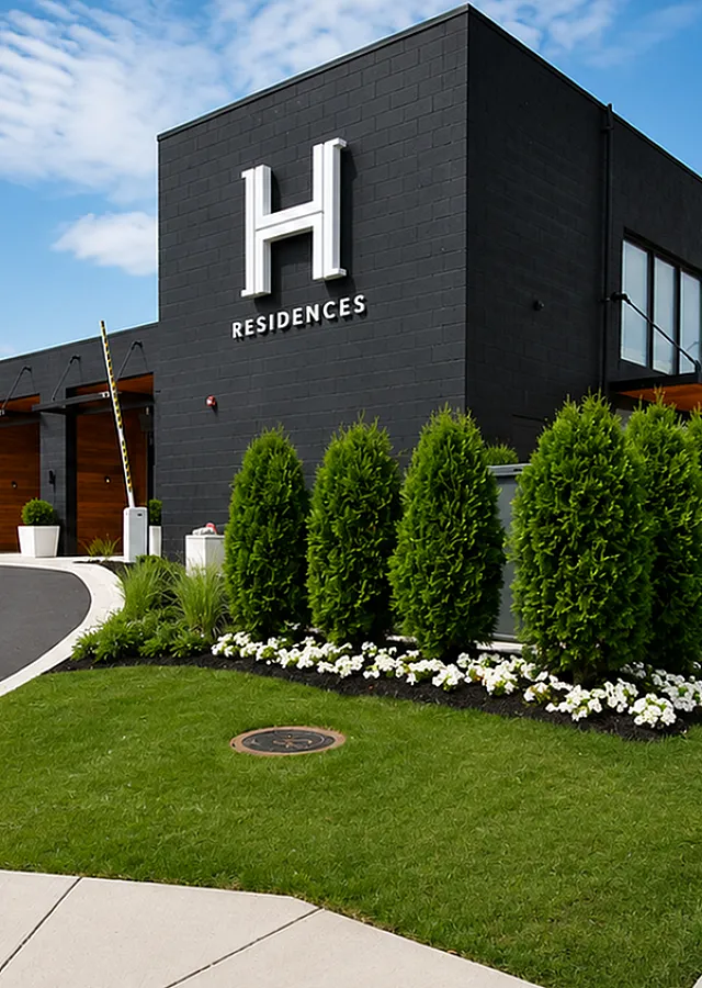 Modern apartment complex with sleek black exterior, stylish wooden accents, and lush landscaped gardens under a bright blue sky.