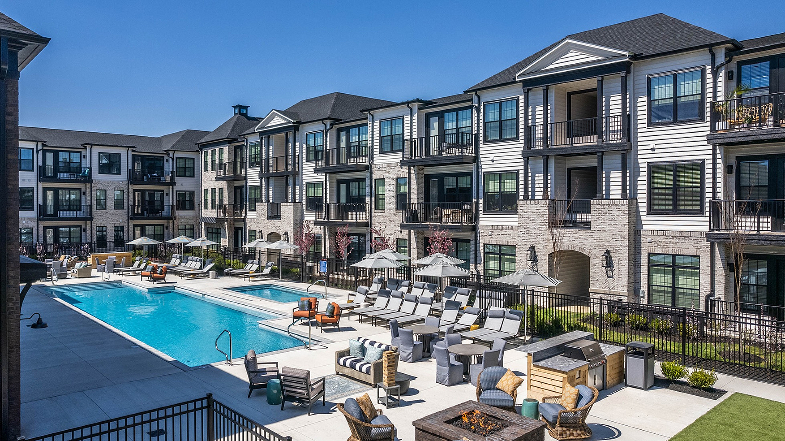 Luxurious apartment complex with a spacious swimming pool, stylish outdoor lounge, and modern balconies under a clear blue sky.
