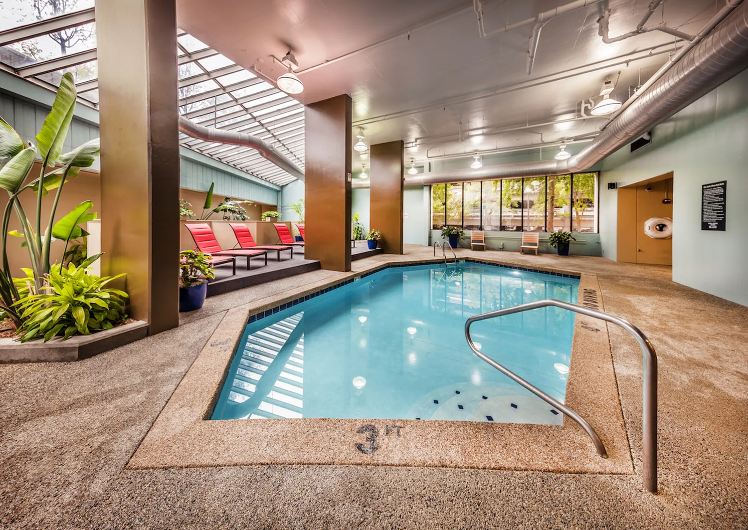 Inviting indoor pool area with sleek skylight, vibrant red lounge chairs, and lush greenery for ultimate relaxation.