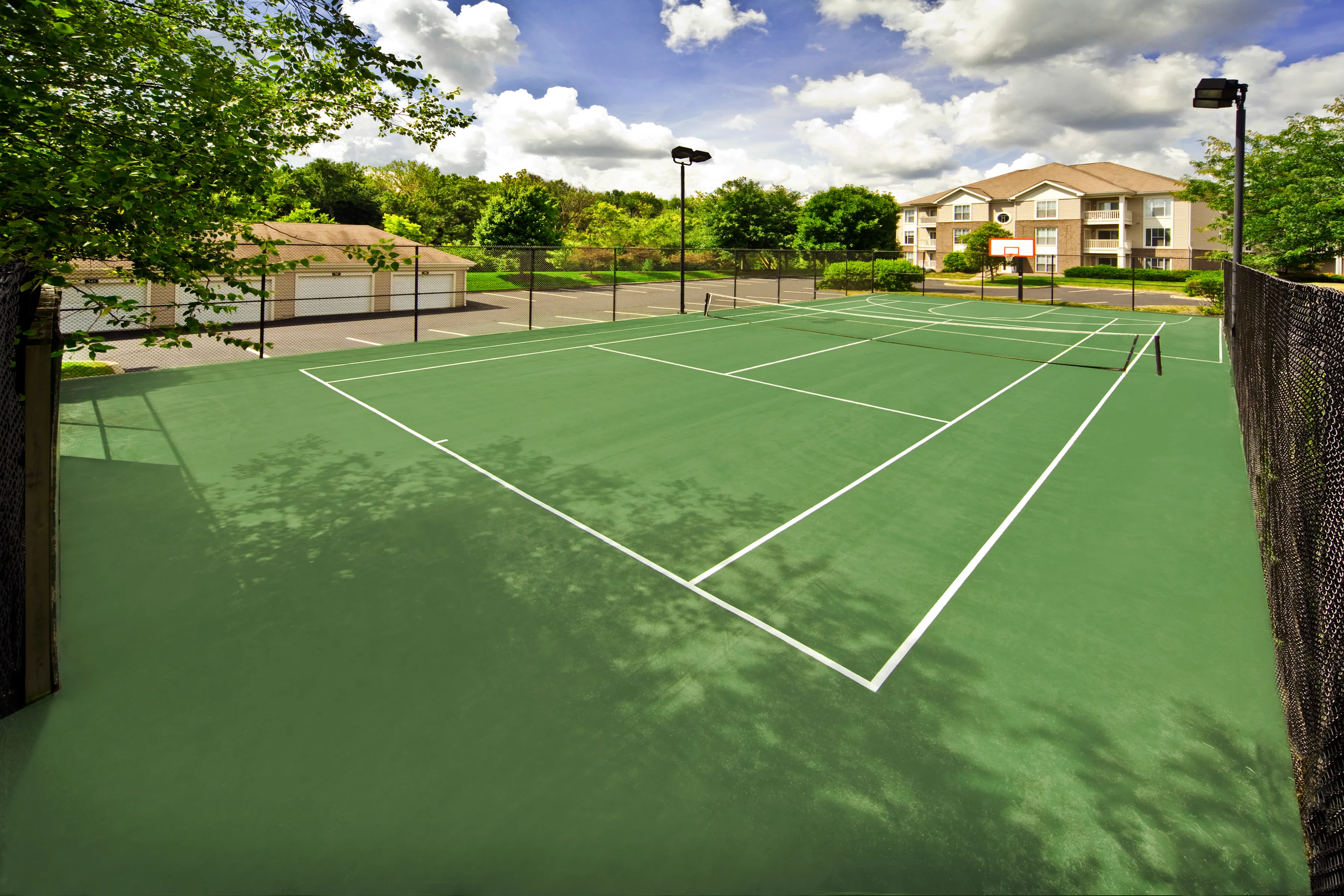 Scenic apartment community with pristine tennis court, surrounded by lush greenery and charming residences in the background.