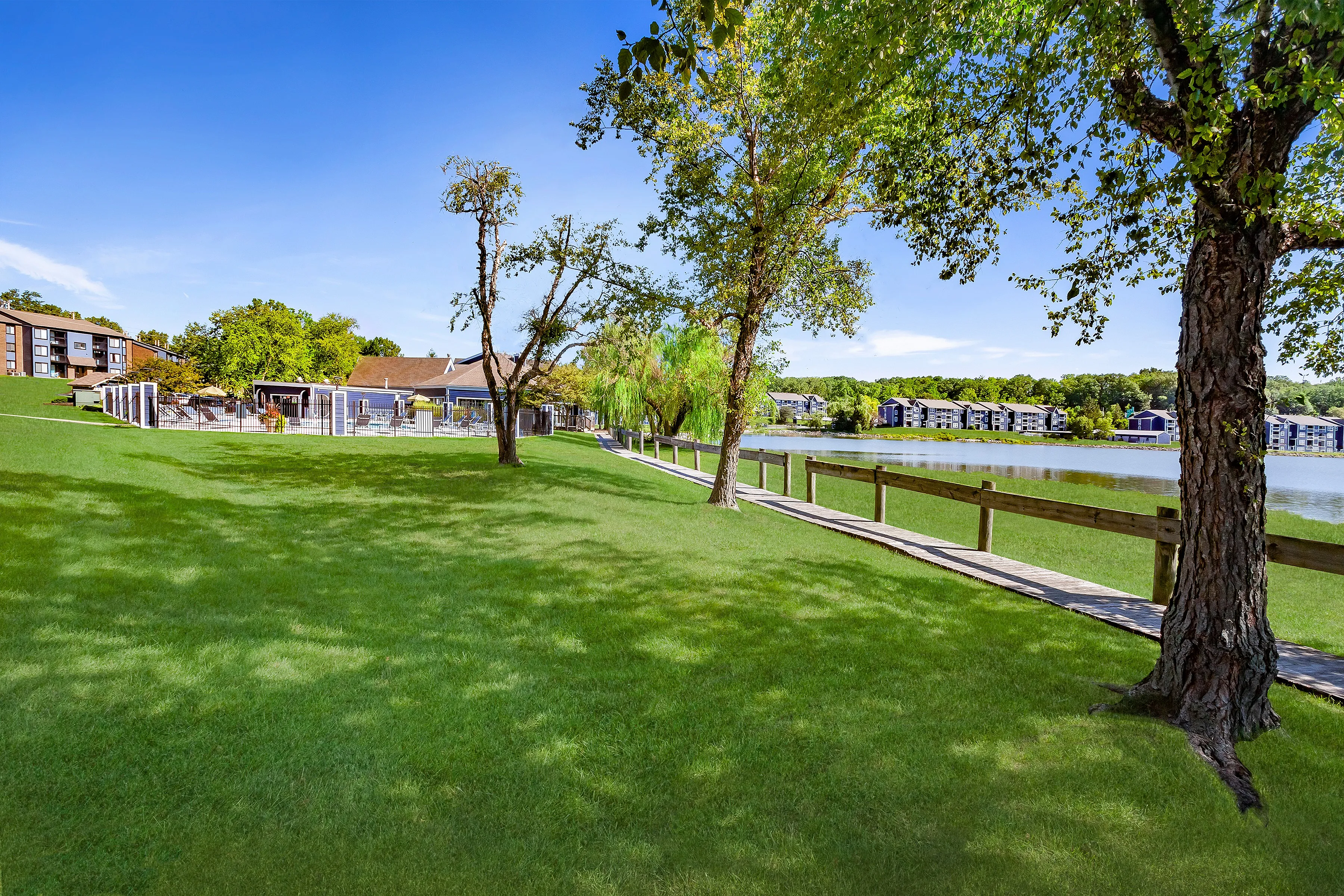 Serene lakefront area with lush green lawns, a boardwalk, and modern apartment buildings.