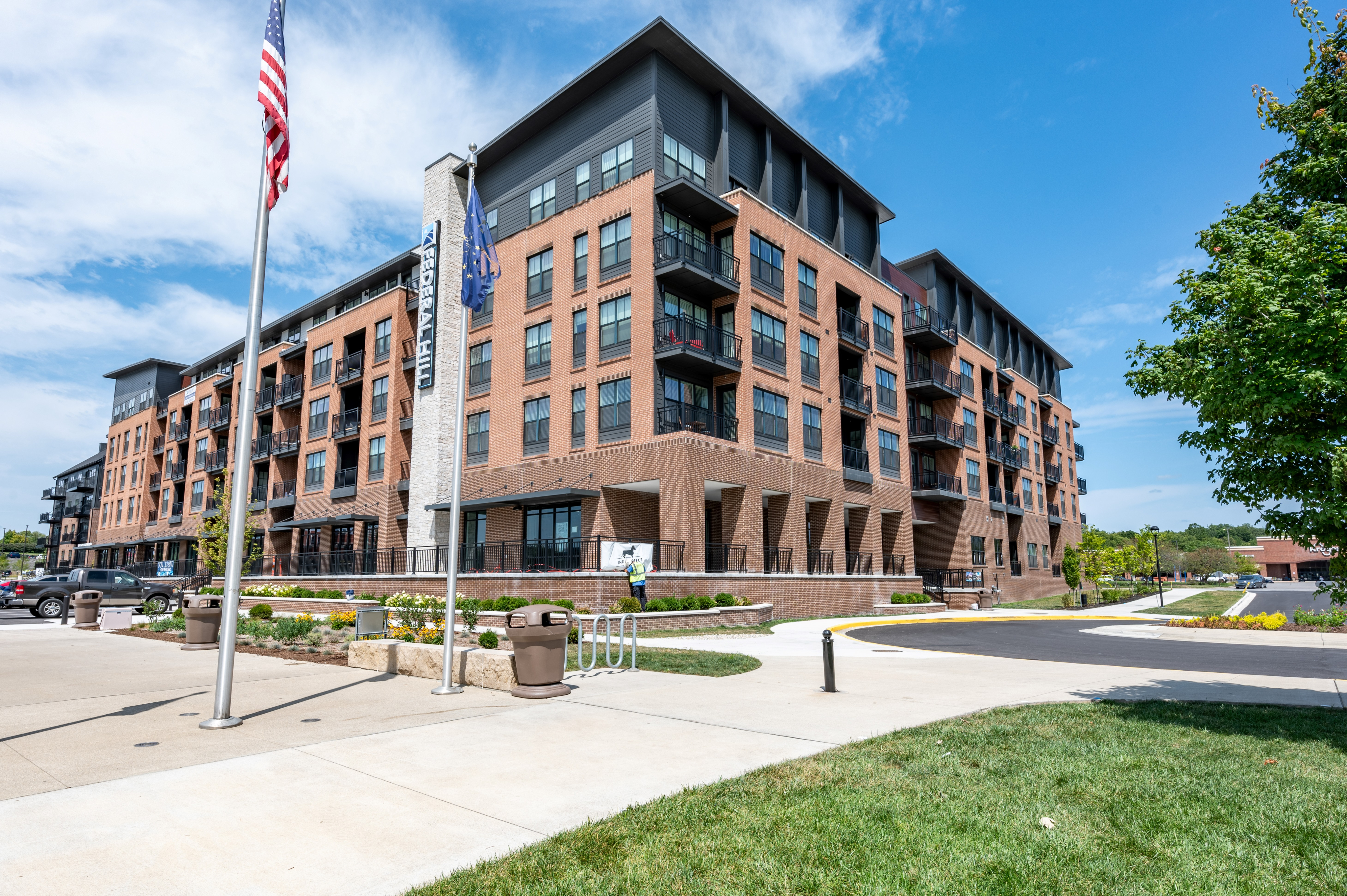 Modern apartment building with stylish brick facade, large windows, and spacious balconies, set against a clear blue sky, offering urban living with attractive landscaping.